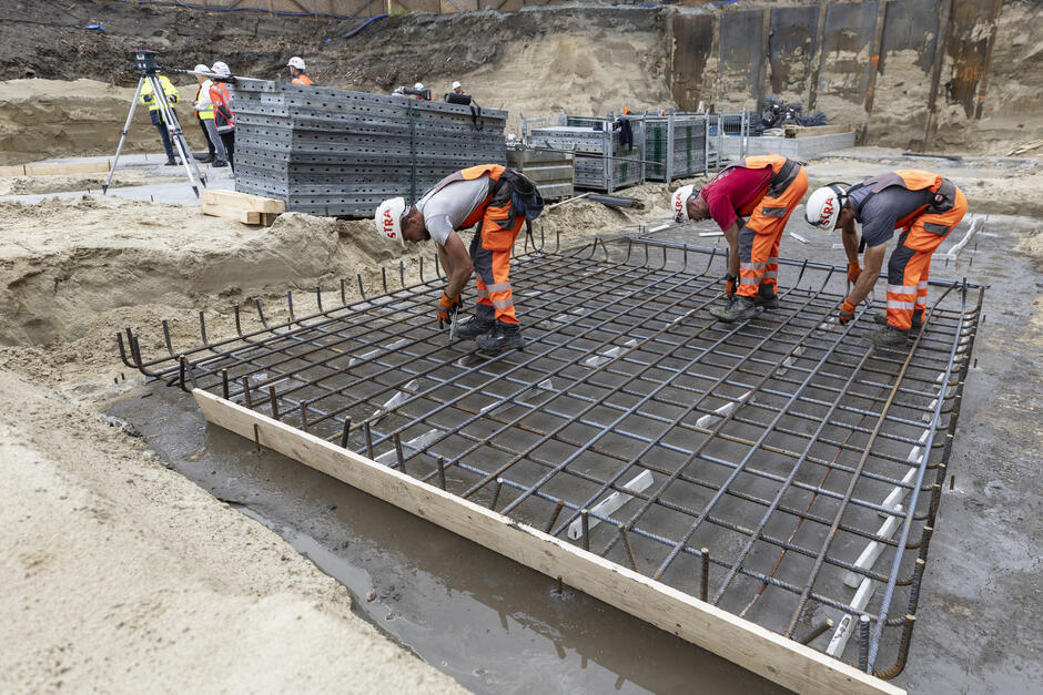 na zdjęciu trzech robotników w roboczych pomarańczowych kombinezonach, układają dużą kratę na wylanym w ziemi betonie