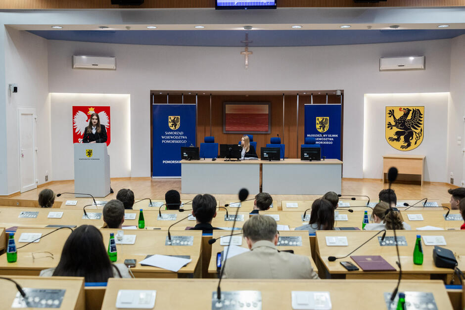 Sesja Młodzieżowego Sejmiku Województwa Pomorskiego. W sali plenarnej siedzą młodzi ludzie, widoczni od tyłu, na mównicy przemawia młoda kobieta zwrócona do siedzących w ławach, za długim stołem siedzi inna młoda kobieta. Po bokach widać herby Polski i województwa pomorskiego.