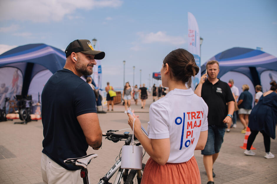 Kobieta w koszulce z napisem „Trzymaj pion” rozmawia z mężczyzną prowadzącym rower na tle namiotów promocyjnych.