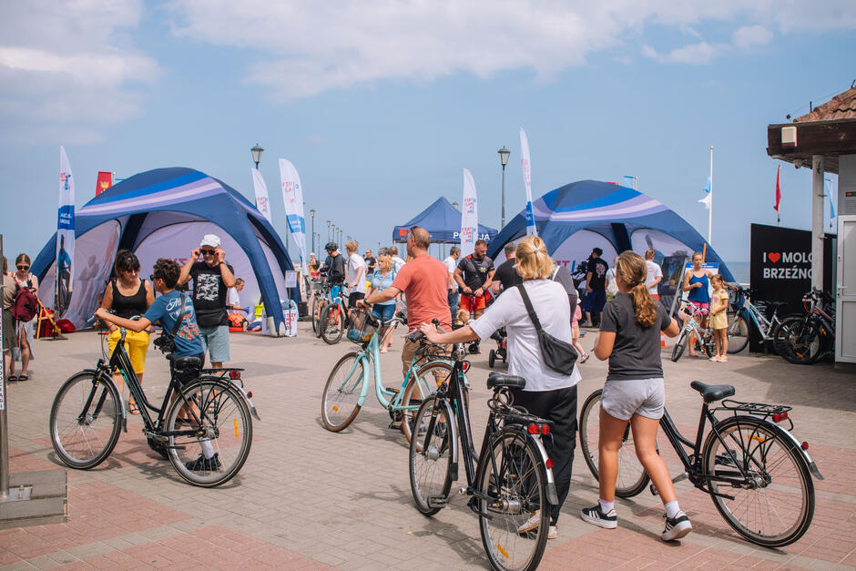 Tłum ludzi z rowerami spaceruje deptakiem obok niebieskich namiotów kampanii edukacyjnej przy plaży w Brzeźnie.