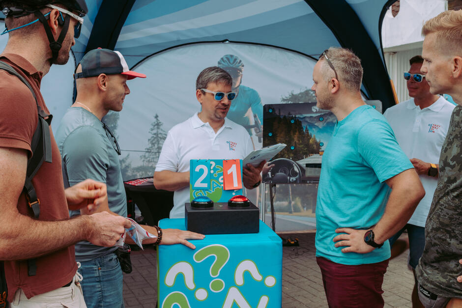 Mężczyźni biorą udział w quizie przy kolorowym stanowisku z przyciskami i tablicą wyników w namiocie kampanii „Trzymaj pion”.