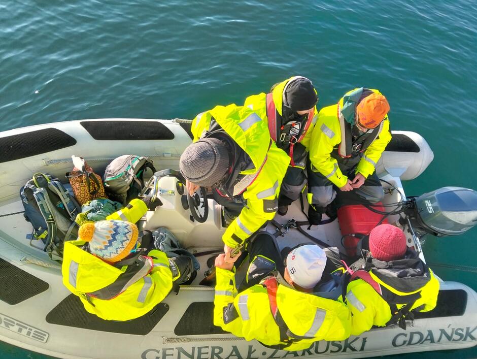 Na zdjęciu widoczna jest grupa osób w jaskrawożółtych kurtkach asekuracyjnych siedzących w pontonie motorowym na spokojnej wodzie. Wygląda na to, że przygotowują się do lądowania lub powrotu z wyprawy, co sugeruje ich ubiór oraz sprzęt turystyczny na pokładzie. 