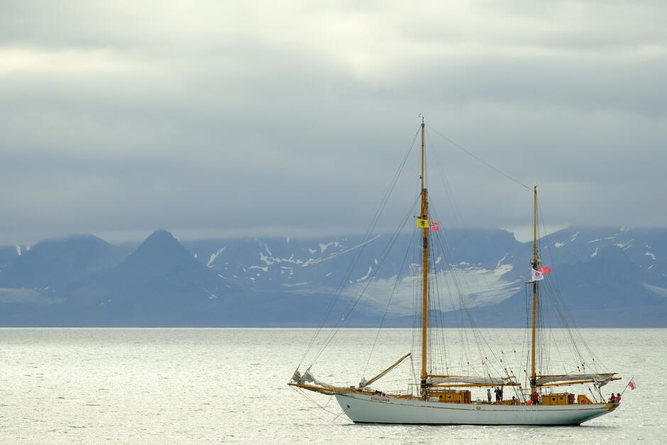 Na zdjęciu widoczny jest żaglowiec spokojnie unoszący się na wodach arktycznego fiordu, z majestatycznymi, ośnieżonymi górami w tle. Ponure, niskie chmury nadają scenerii surowy, tajemniczy klimat, typowy dla rejonów Svalbardu.
