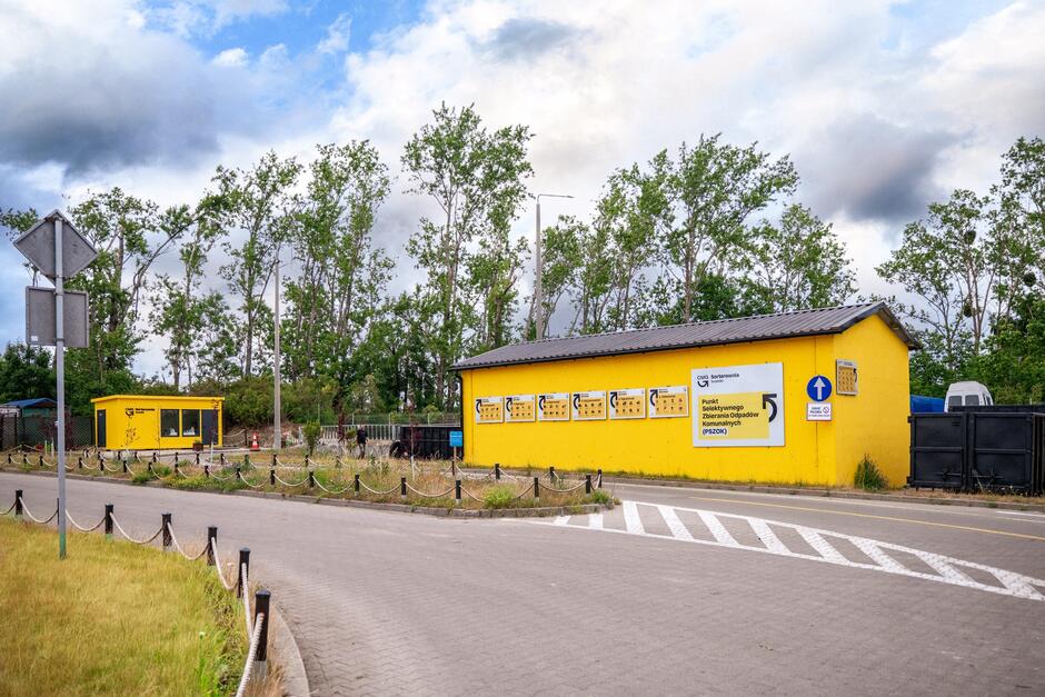 Pomalowane na żółto budynki punktu odbioru odpadów. W tle rosną drzewa.