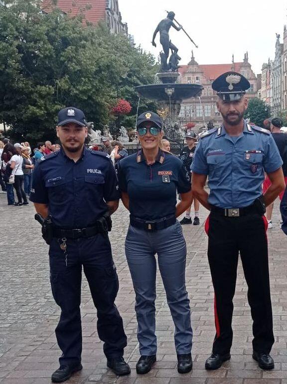 na zdjęciu dwóch policjantów i jedna policjantka w mundurach, pozują do zdjęcia, za nimi widać fontannę neptuna