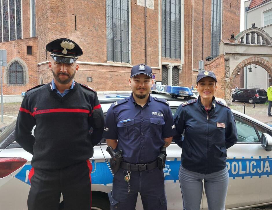 na zdjęciu dwóch policjantów i jedna policjantka w mundurach, pozują do zdjęcia, za nimi stoi radiowóz