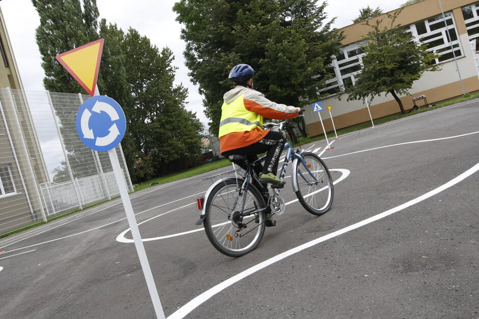 na zdjęciu dziecko jadące na rowerze, widać je od tyłu, w kasku, jedzie po miasteczku ruchu drogowego