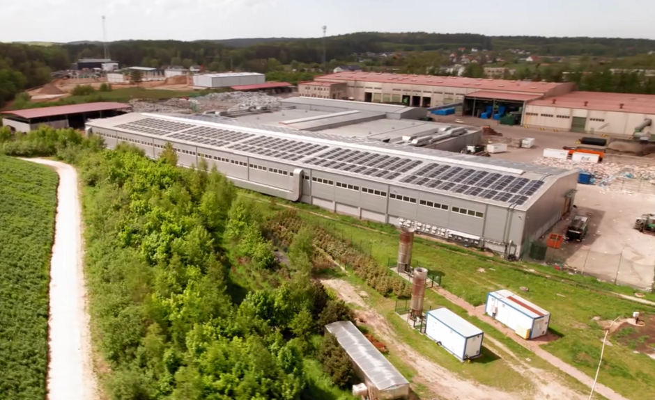 Długi budynek z dachem pokrytym panelami słonecznymi. Wokół rosną drzewa.