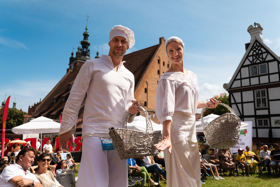 Na zdjęciu widzimy kobietę i mężczyznę ubranych w tradycyjne białe stroje, którzy uśmiechają się do obiektywu, trzymając wiklinowe kosze. W tle znajduje się malowniczy rynek z zabytkowymi budynkami oraz grupa obserwujących wydarzenie ludzi, co sugeruje, że trwa festyn lub lokalne święto.
