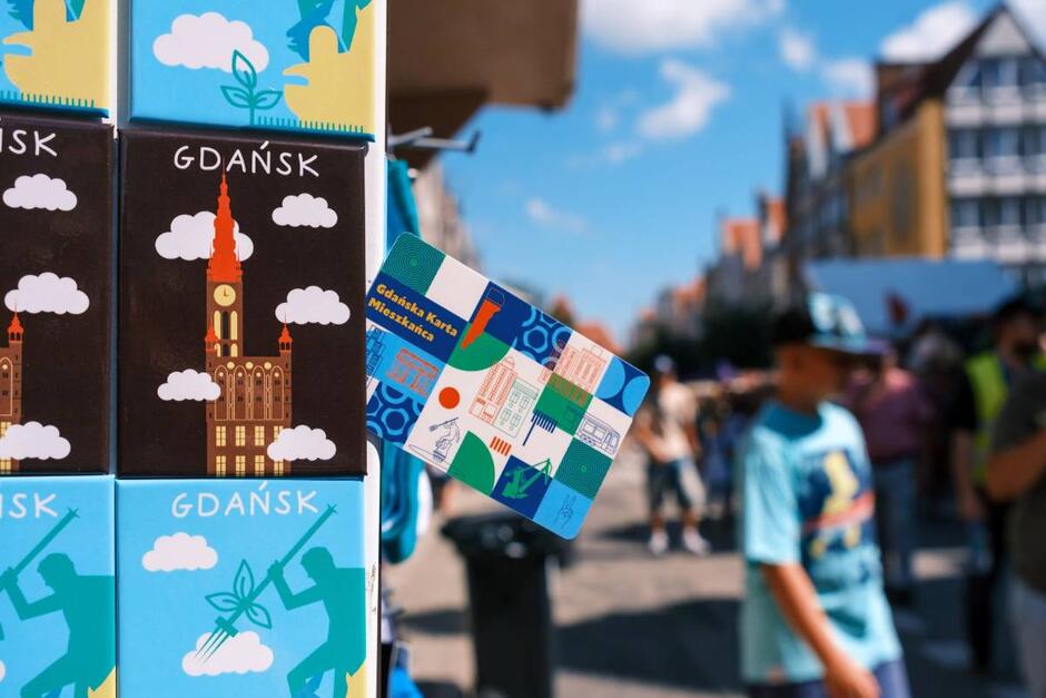 Na zdjęciu widać kolorowe pamiątki z Gdańska oraz Gdańską Kartę Mieszkańca  umieszczone na wystawie stoiska podczas słonecznego dnia. W tle rozmyty tłum ludzi spacerujących po deptaku wśród zabytkowych kamienic tworzy atmosferę letniego jarmarku lub festynu.
