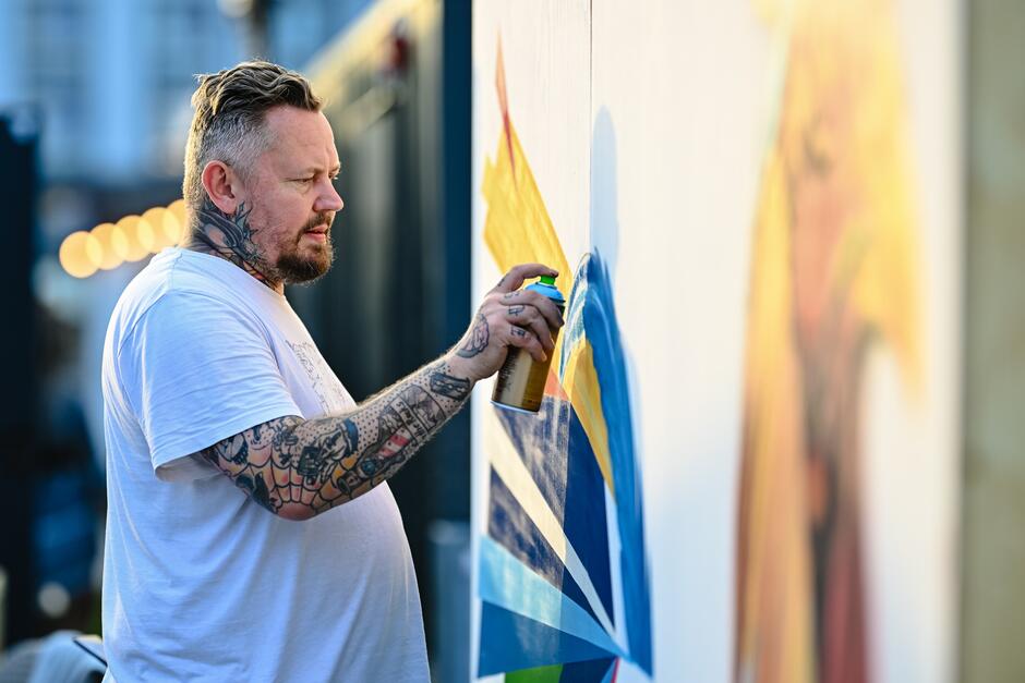 Mężczyzna maluje sprayem na ścianie koguta. Na szyi i ręku ma tatuaże.