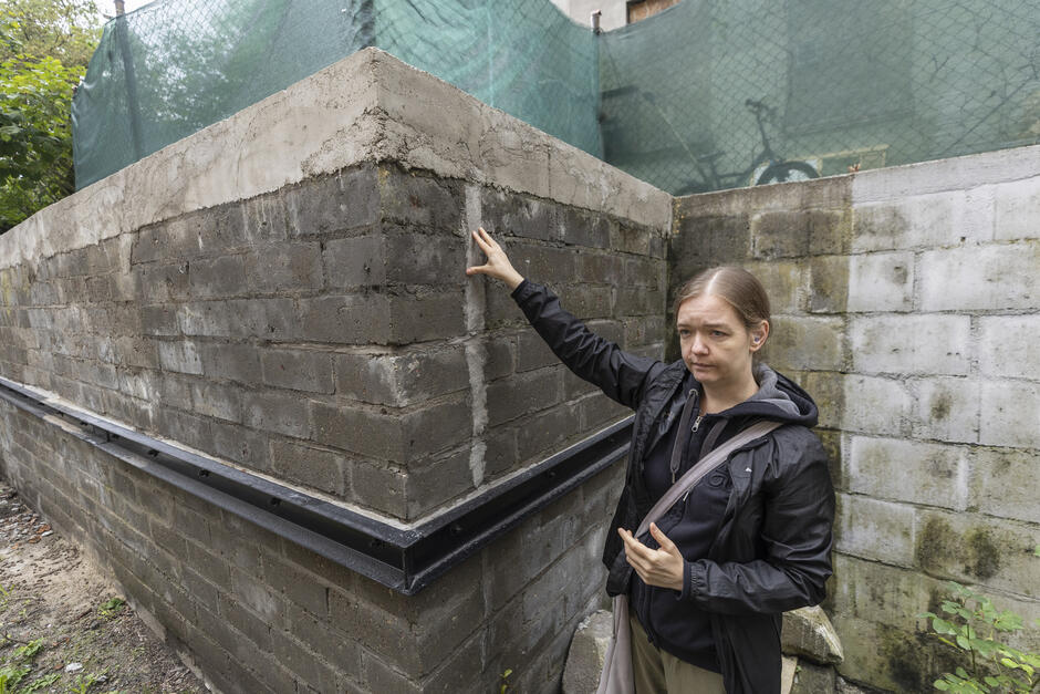 na zdjęciu kobieta w średnim wieku stoi przy szarym ceglanym murze oporowym