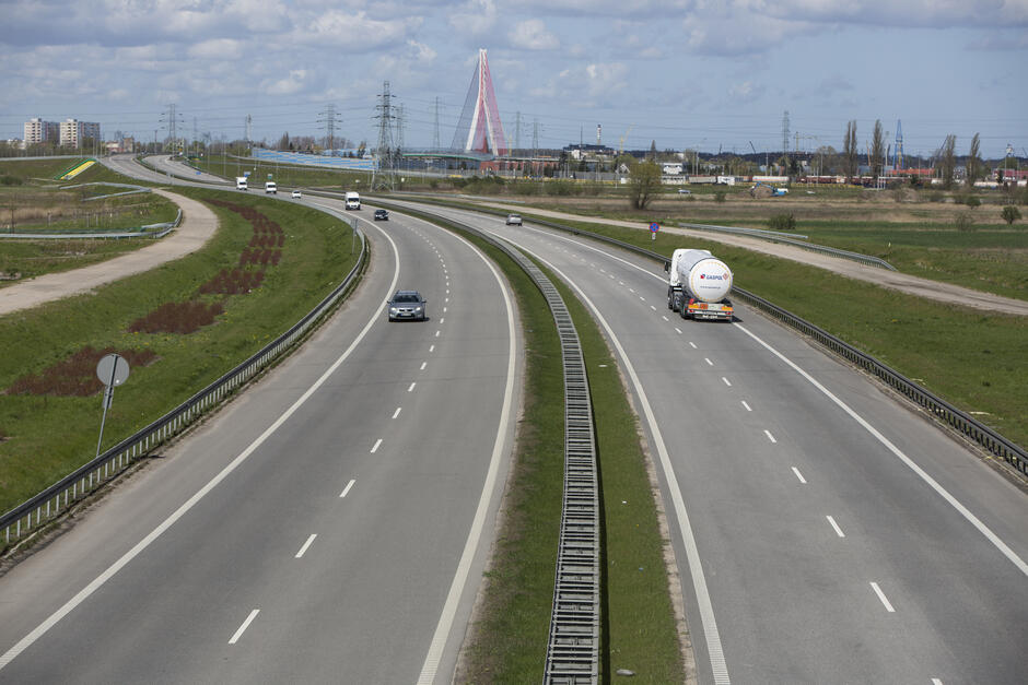 Na zdjęciu widać szeroką, wielopasmową drogę szybkiego ruchu, biegnącą przez otwartą, zieloną przestrzeń. Droga jest podzielona pasem zieleni z barierkami ochronnymi pośrodku. Ruch jest umiarkowany – widać kilka samochodów osobowych i jeden większy pojazd ciężarowy-cysterna, jadący w kierunku prawej strony zdjęcia. W tle rozciąga się panorama miasta z zabudowaniami przemysłowymi, liniami wysokiego napięcia oraz charakterystycznym mostem o czerwonej konstrukcji linowej. Niebo jest częściowo zachmurzone, a światło wskazuje na pogodny dzień.