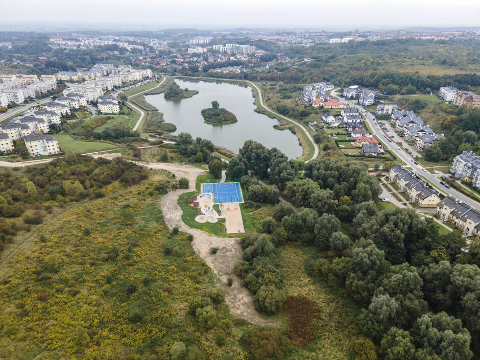 Na zdjęciu z lotu ptaka widać rozległy, zielony teren rekreacyjny z dużym stawem o wydłużonym kształcie i niewielką wyspą pośrodku. Wokół zbiornika wodnego poprowadzone są ścieżki spacerowe, a brzegi porastają drzewa i krzewy.