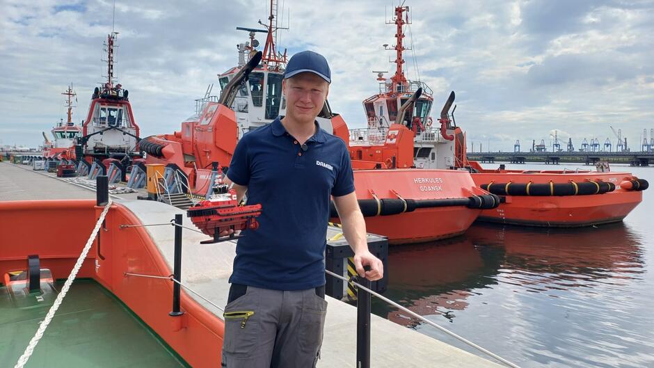 Na zdjęciu widać mężczyznę stojącego na nabrzeżu portowym, trzymającego w ręku model małego statku w kolorze czerwonym. Ubrany jest w granatową koszulkę polo z logo „DAMEN” oraz czapkę z daszkiem, a także szare spodnie robocze z zielonym suwakiem w kieszeni.