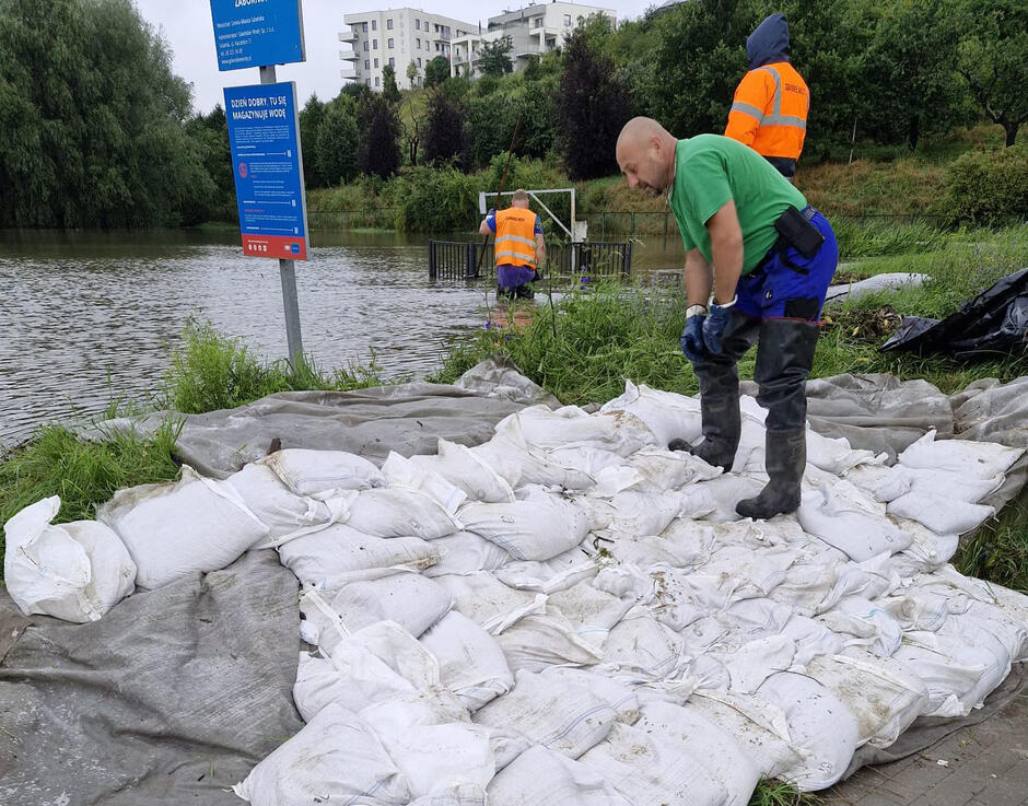 Staw retencyjny, ba brzegu mężczyzna układa worki z piaskiem. 
