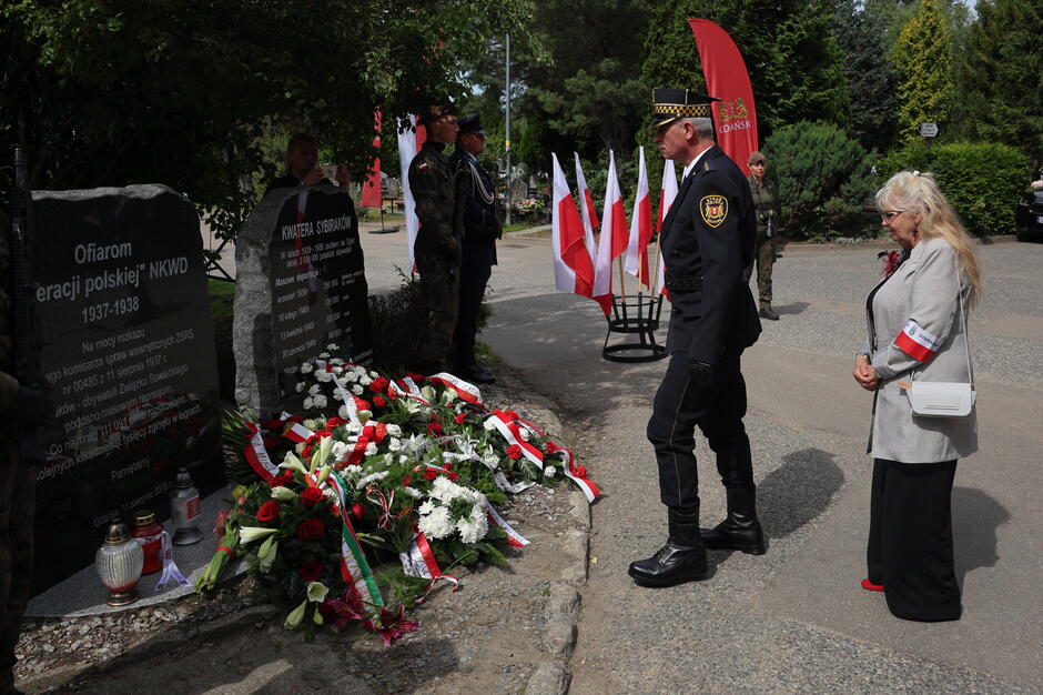 na zdjęciu kobieta stoi przed pomnikiem, a pod nim leży dużo wiązanek kwiatów, obok kobiety stoi strażnik miejski w mundurze