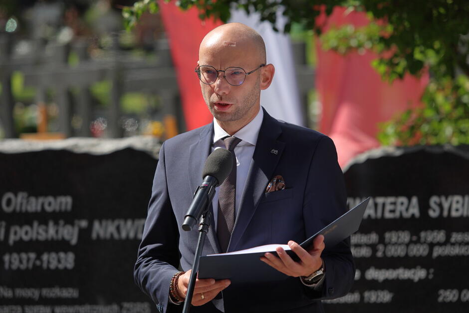 na zdjęciu mężczyzna w średnim wieku, w garniturze, trzyma kartkę, mówi do mikrofonu