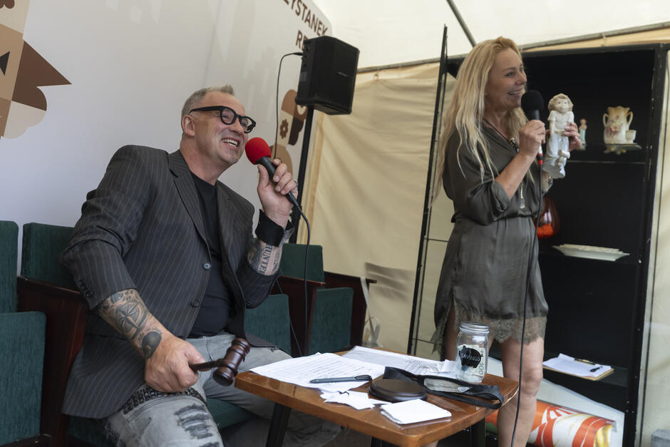 Na zdjęciu widzimy scenę z aukcji lub licytacji. Po lewej stronie siedzi mężczyzna w ciemnej marynarce w prążki, czarnej koszulce i jasnych spodniach z przetarciami. Ma krótkie, siwe włosy, nosi czarne okulary i ma liczne tatuaże na przedramionach. W jednej ręce trzyma młotek aukcyjny, a w drugiej mikrofon z czerwoną gąbką, uśmiechając się szeroko.