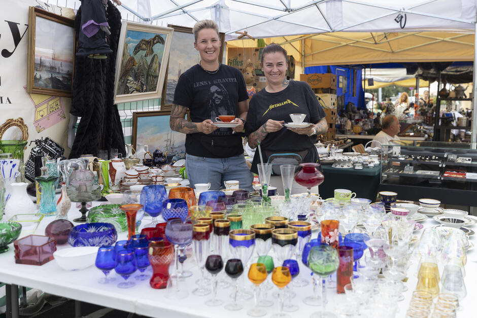 Na zdjęciu widać dwie uśmiechnięte kobiety stojące za stoiskiem na targu lub jarmarku, prawdopodobnie sprzedające antyki lub przedmioty vintage. Obie trzymają w rękach filiżanki z talerzykami. Stół przed nimi jest pełen kolorowego szkła – kieliszków, pucharów i wazonów w różnych kształtach i barwach, od zielonego, przez czerwony i pomarańczowy, po fioletowy i kobaltowy.