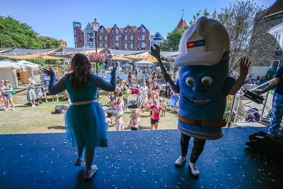 Na zdjęciu widać scenę, na której stoją dwie postacie – po lewej kobieta w turkusowej sukience z tiulem i długimi rękawami, a po prawej osoba w dużym, niebieskim kostiumie maskotki z kaskiem ochronnym z logo „GIWK”. Maskotka ma zabawny, lekko groźny wyraz twarzy i szeroki uśmiech z zębami. Patrząc z perspektywy sceny, w tle widzimy publiczność – głównie dzieci, ale także kilku dorosłych – stojącą i siedzącą na trawie. Ludzie wydają się zaangażowani i uśmiechnięci, niektóre dzieci machają rękami lub biegają.