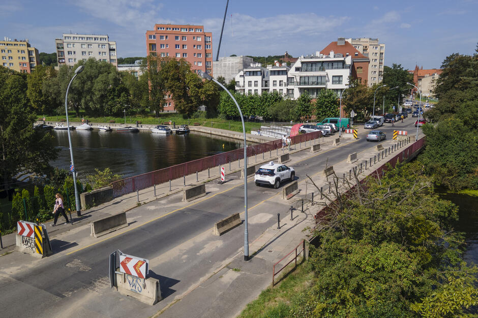 zdjęcie z drona, widać most po którym jedzie samochód a obok przechodzi kobieta, w tle budynki