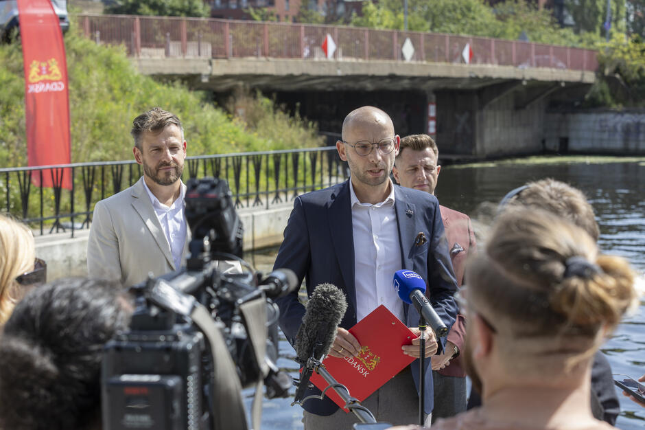 na zdjęciu mężczyzna w okularach łysy mówi do dziennikarskich mikrofonów, za nim stoją dwaj mężczyźni w średnim wieku