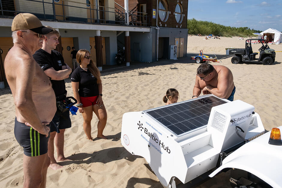Wokół robota sprzątającego plażę zebrało się kilka osób, do pojemnika na śmieci w środku robota zagląda mała dziewczynka.