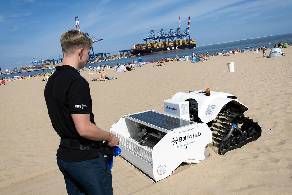 Mężczyzna idzie po plaży sterując jadącym obok robotem, który czyści plażę. W tle plażowicze, morze oraz port.