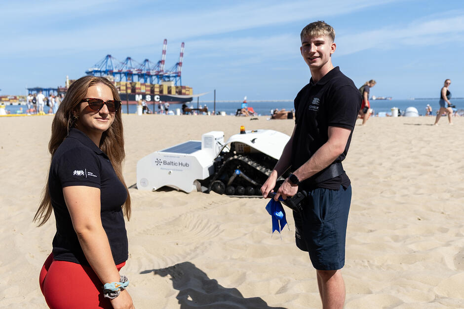 Młoda kobieta i młody mężczyzna pozują do zdjęcia na plaży, za nimi stoi na piasku robot do sprzątania plaży. 