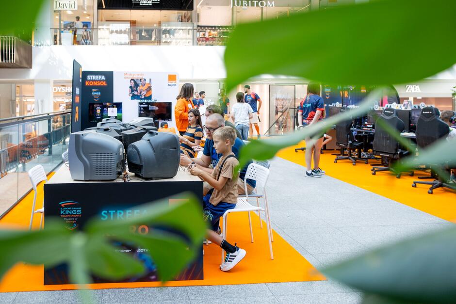 Strefa gier komputerowych w centrum handlowym. Przed komputerem siedzi dorosły mężczyzna z chłopcem i grają.