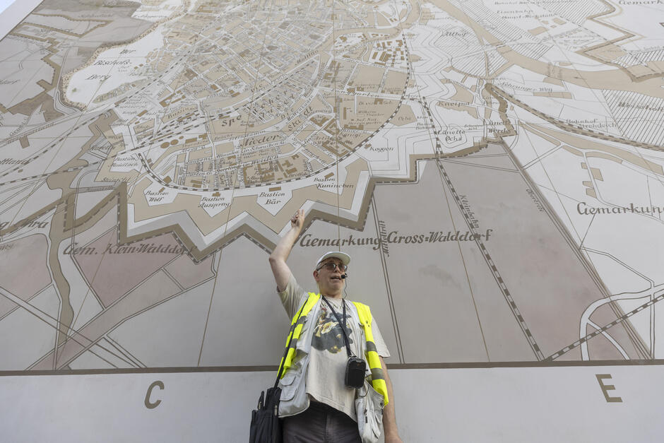 mężczyzna w jaskrawej kamizelce pozuje na tle muralu na ścianie, mural przedstawia starą mapę