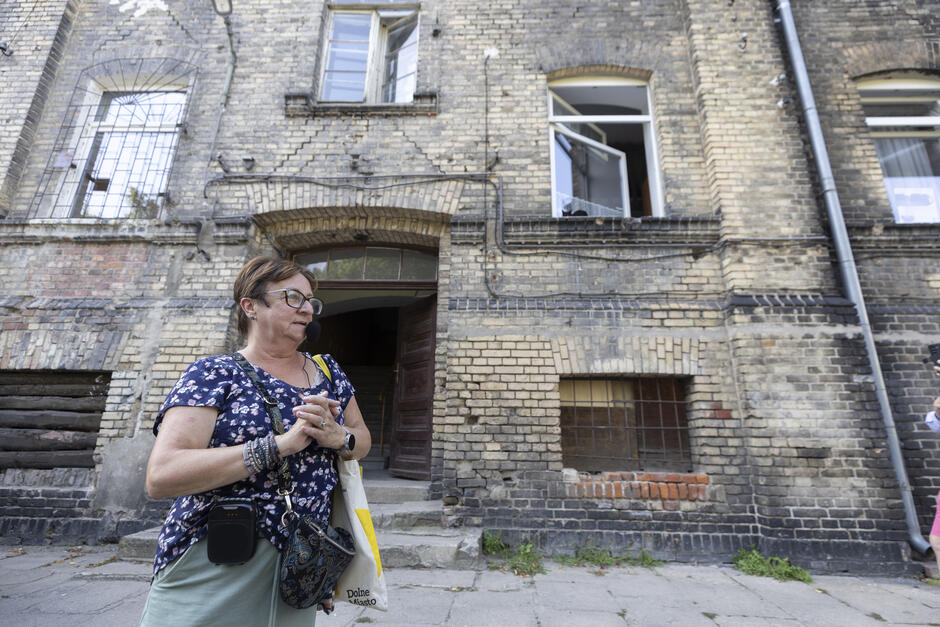na zdjęciu dojrzała kobieta stoi przy starym zabytkowym ceglanym budynku