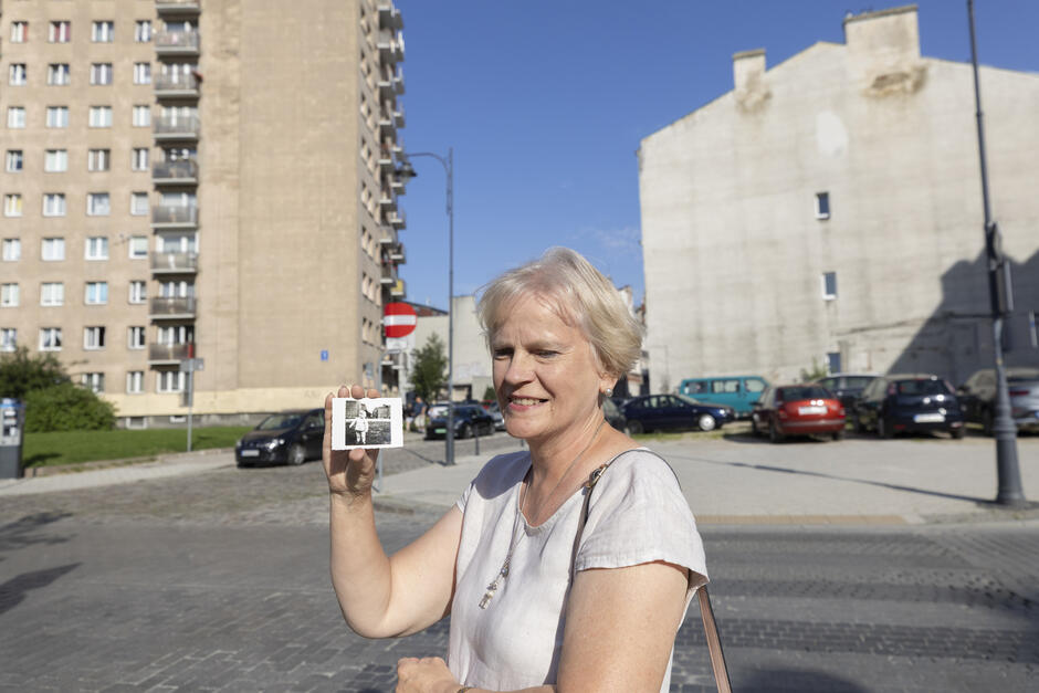 na zdjęciu dojrzała kobieta trzyma w ręku fotografię, za nią stoją dwa wysokie budynki