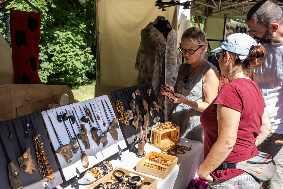 Stragan z wyrobami z drewna, bursztynu i muszli przy którym stoją trzy osoby.