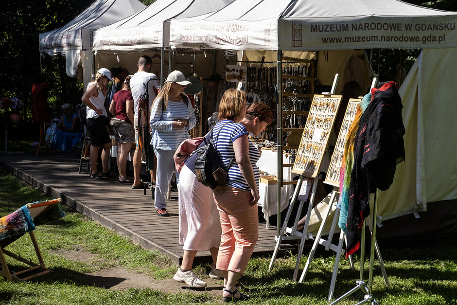 Stragany z rękodziełem na jarmarku w ogrodzie, przed straganami ludzie.