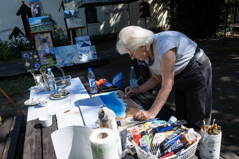Starsza kobieta maluje obraz na stole ustawionym w ogrodzie. Za nią obrazy na sprzedaż.