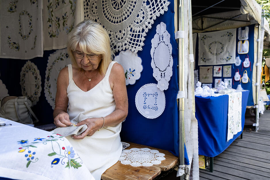 Starsza kobieta haftuje na straganie na jarmarku.
