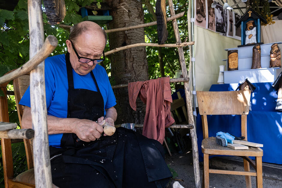 Starszy mężczyzna rzeźbi w drewnie ptaka siedząc na krześle w ogrodzie, obok stoi jego stragan na jarmarku z rzeźbami.