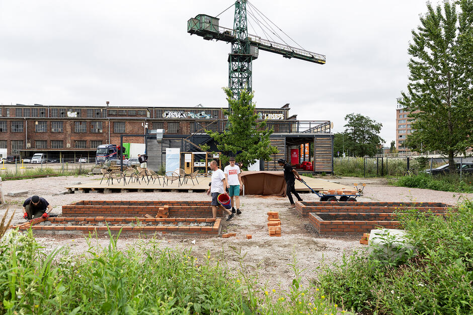 Kilku mężczyzn pracuje robiąc ceglane murki, za którymi mają być grządki. Wokół nich przestrzeń przemysłowa - hala i dźwig stoczniowy.