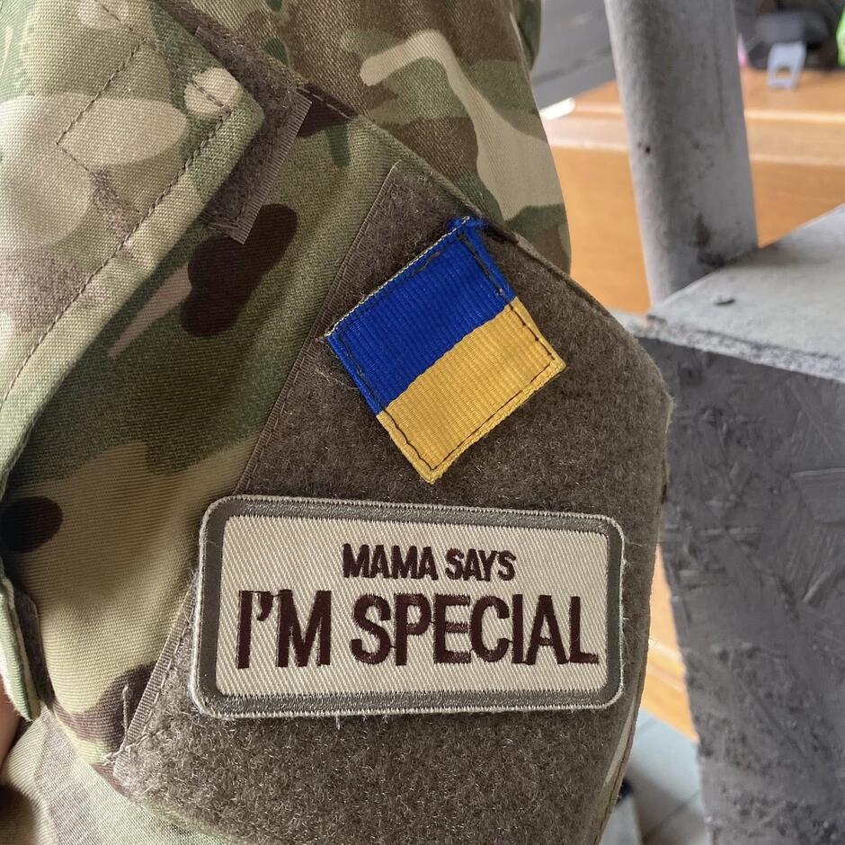 Ramię żołnierza ubranego w moro, na nim dwie naszywki. U góry w kwadracie flaga Ukrainy, poniżej napis Mama says I'm special