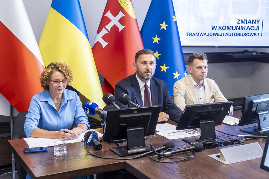 na zdjęciu przy stole siedzą dwa mężczyźni i jedna kobieta, ta mówi do mikrofonów dziennikarskich