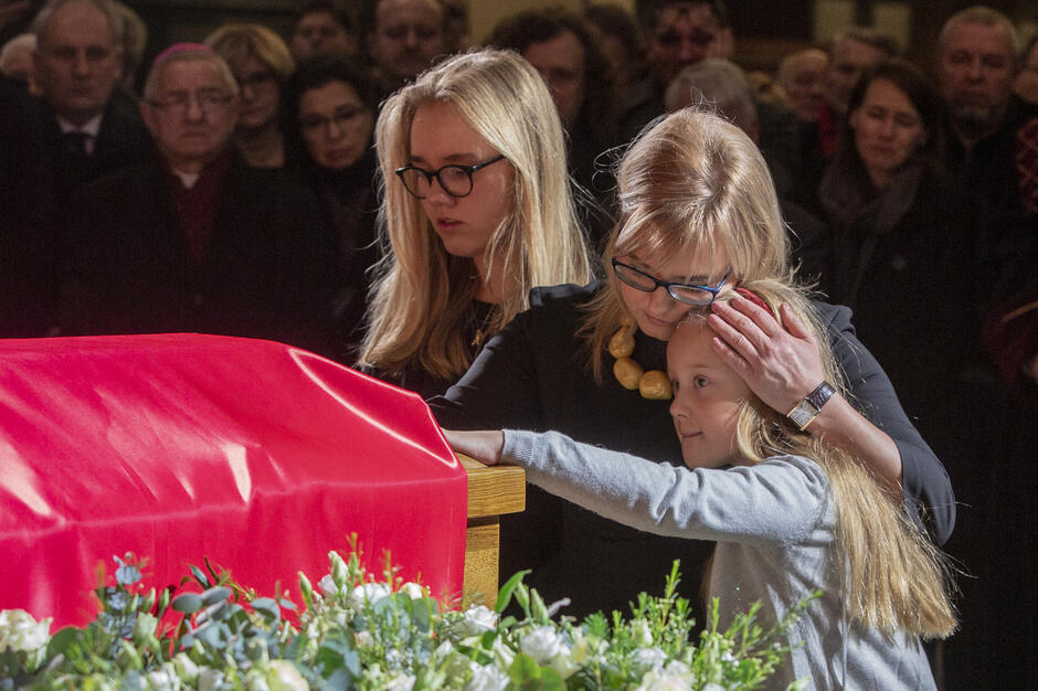 Kobieta z dwójką przytulonych dziewczyn stoją przed trumną podczas uroczystości pogrzebowych.