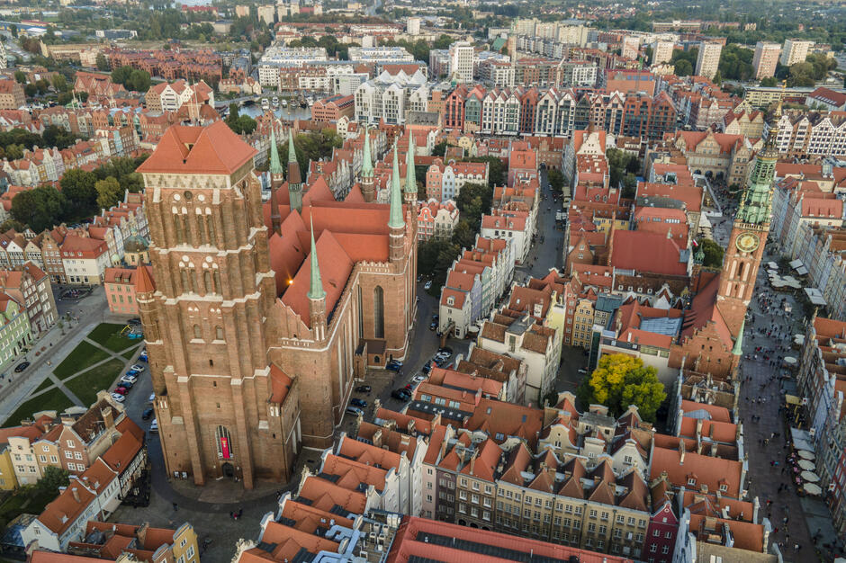Widok z lotu ptaka na historyczne centrum Gdańska. W centrum zdjęcia znajduje się Bazylika Mariacka z czerwonym dachem i charakterystycznymi wieżami. Po prawej stronie widoczny jest Ratusz Głównego Miasta z wysoką wieżą zegarową. Otaczające budynki mają czerwone dachy i różnorodne fasady. W tle widać nowoczesne zabudowania oraz zielone tereny miejskie.