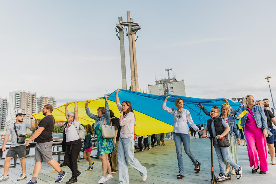 Ludzie maszerują przez miasto i niosą nad głowami wielką ukraińską flagę. W tle pomnik Poległych Stoczniowców w Gdańsku w formie trzech wysokich krzyży.