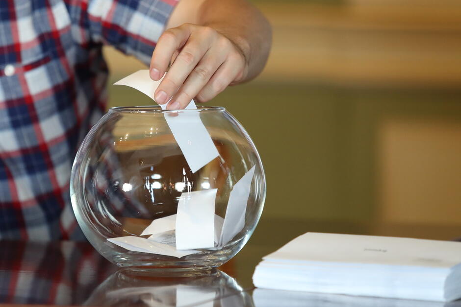 na zdjęciu ludzka ręka wyciąga ze szklanego pojemnika małą, białą kartkę