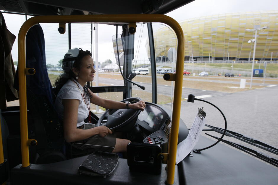 Wnętrze autobusu, za kierownicą młoda kobieta, przez okno widać stadion.