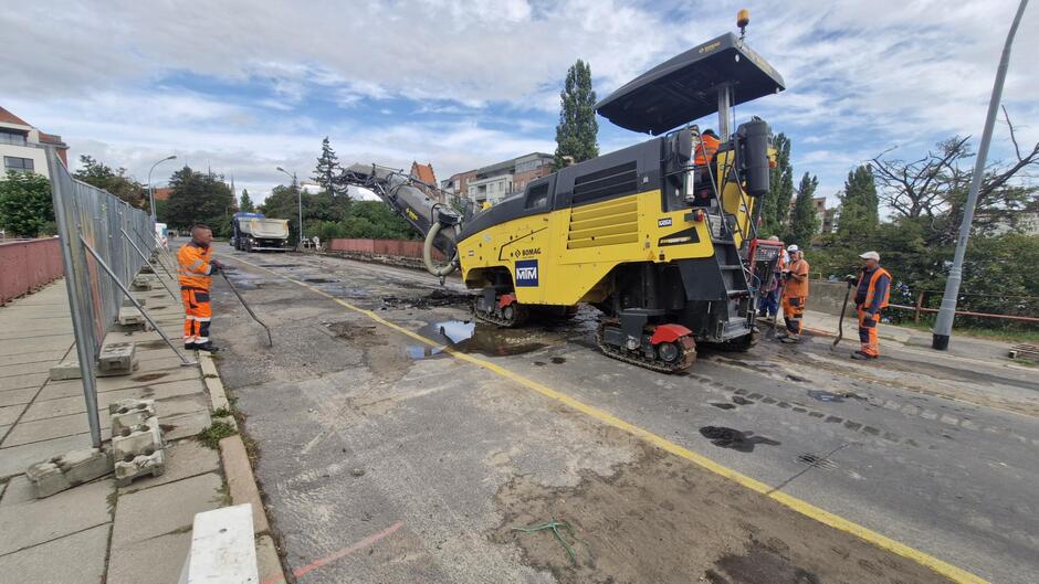 na zdjęciu widać rozbieraną nawierzchnię mostu, po prawej stoi duży pojazd budowlany w żółtym kolorze przy którym stoi kilku robotników