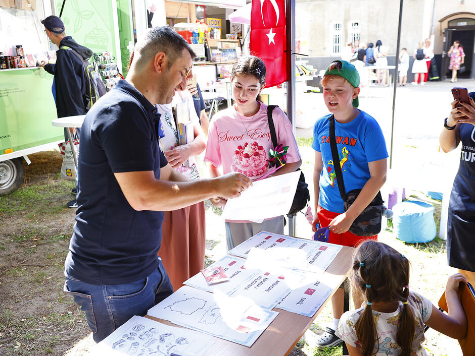 Na straganie z turecką flagą mężczyzna pokazuje coś zgromadzonym wokół dzieciom.
