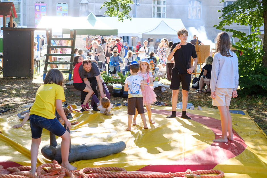 Festyn w ogrodzie w mieście. Na pierwszym planie bawią się dzieci, w tle stragany.